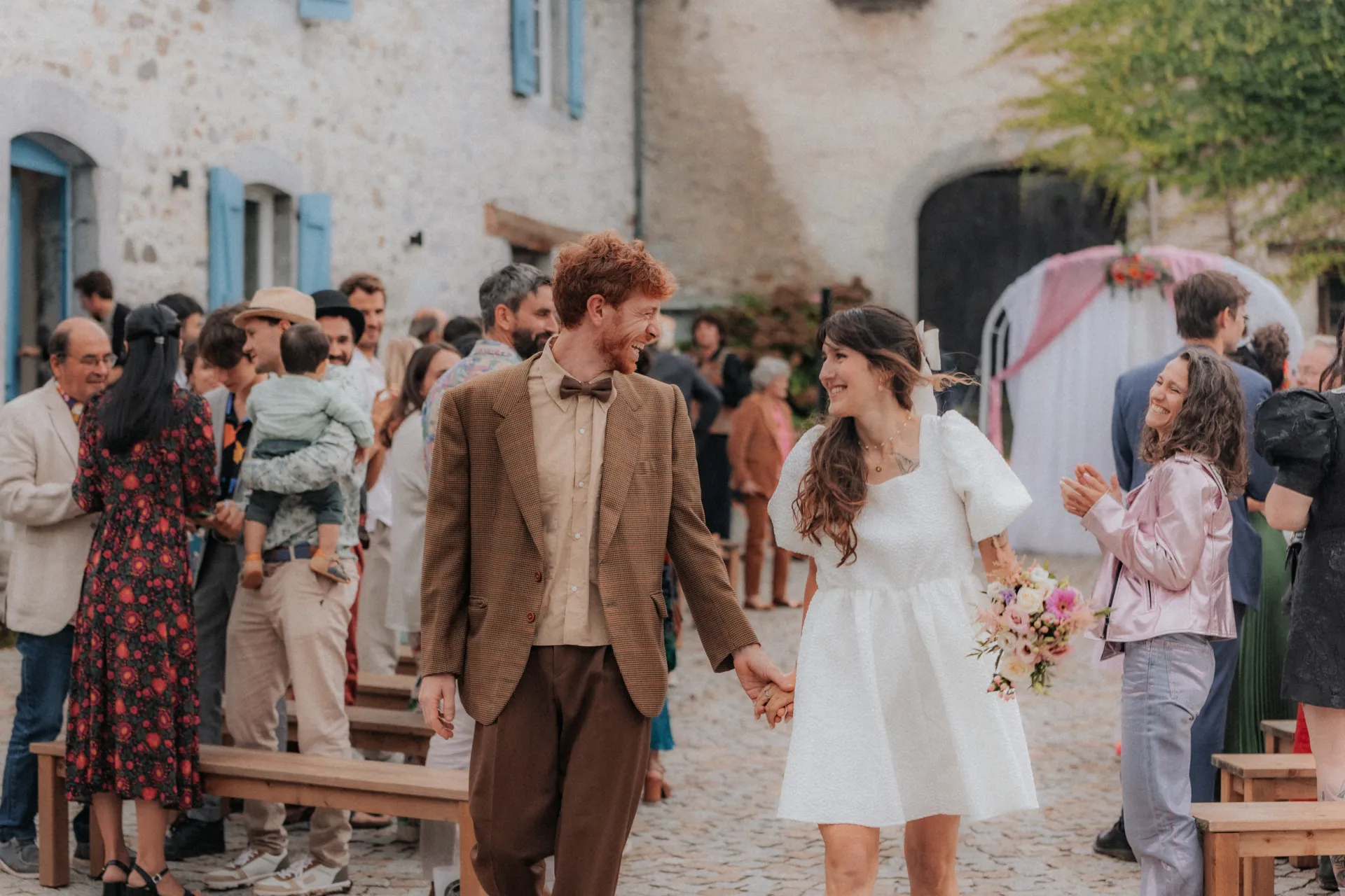 Mariés au style vintage se tenant la main dans une cour en pierre lors d’un mariage nature à Pau