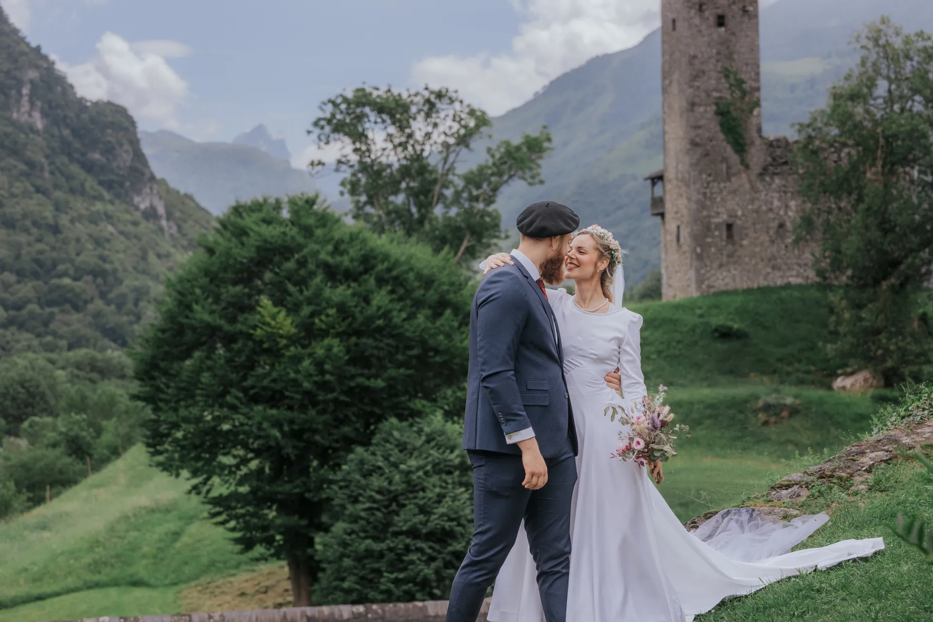 Mariés en séance couple à Castet devant l’Église Saint-Polycarpe, avec montagnes et ruines en arrière-plan