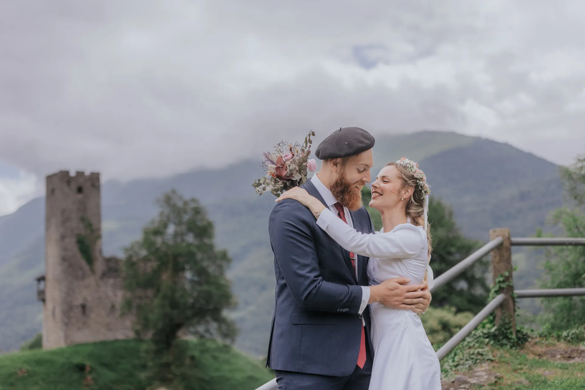 Jeunes mariés enlacés à Castet, près de l’Église Saint-Polycarpe, avec vue sur les montagnes et une tour en arrière-plan