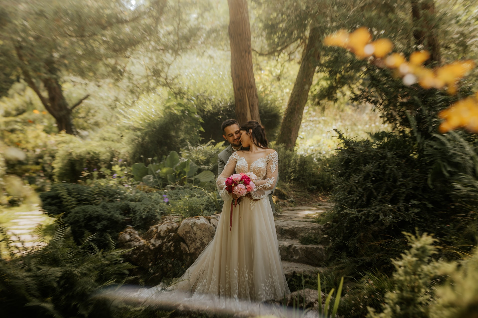 Mariés s'embrassant tendrement dans un jardin verdoyant au Domaine de Bourdale-Dufau, bouquet de fleurs roses en main.