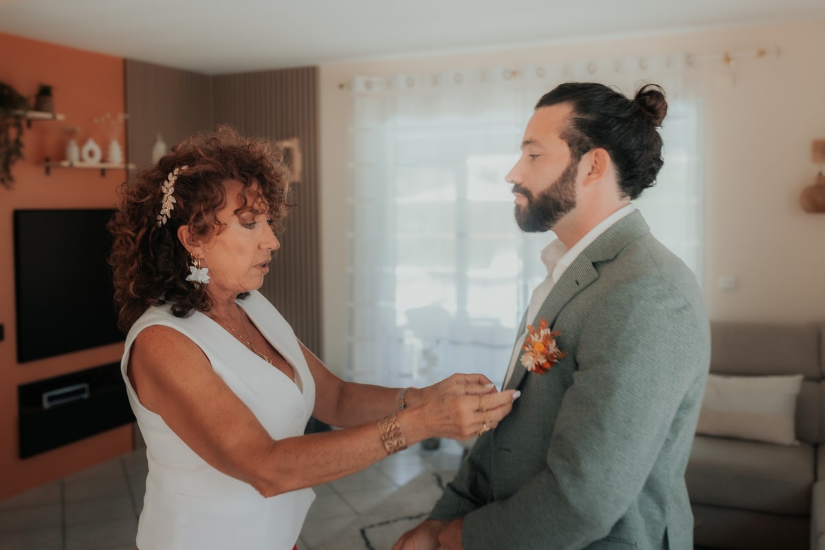 Une femme ajuste la boutonnière d'un homme vêtu d'un costume, dans une maison lumineuse avant un mariage.