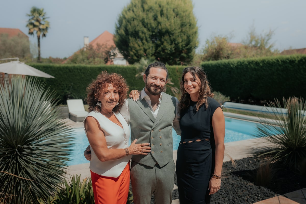 Un homme en costume gris pose avec deux femmes élégantes près d'une piscine, entouré de verdure ensoleillée.