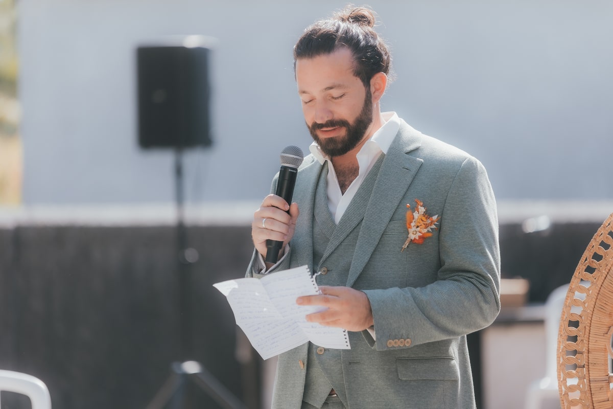 Homme en costume gris lit un discours de mariage, tenant un micro, avec une boutonnière orange, en extérieur à la Villa des 7 Moulins.