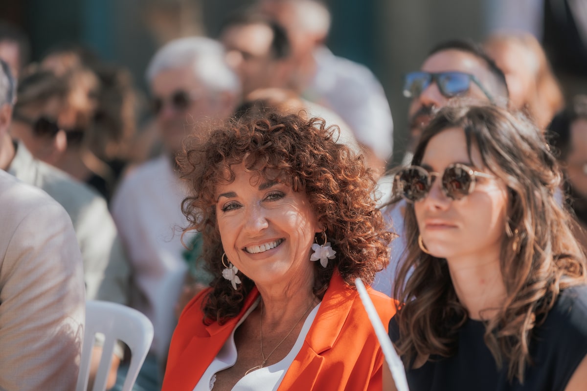 Invitée souriante en veste orange et accessoires floraux, assise parmi les convives du mariage de Manon et Mickael à la Villa des 7 Moulins.