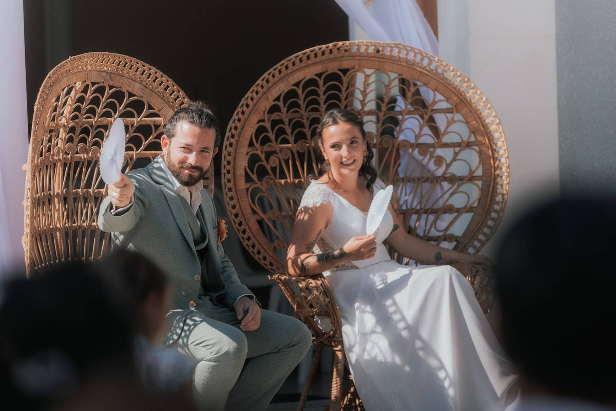 Un couple de mariés assis sur des chaises en rotin, souriant et tenant des éventails lors d'une cérémonie ensoleillée.