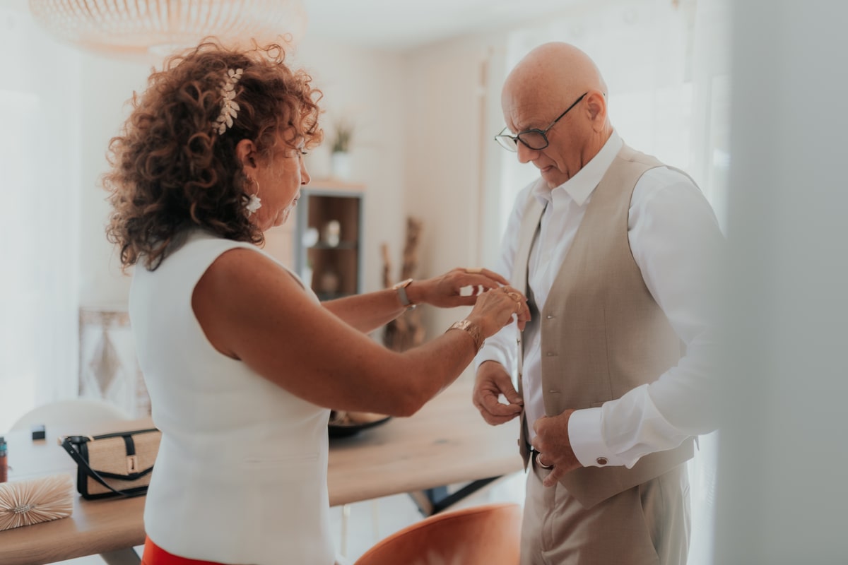 Un homme ajuste sa tenue de mariage avec l'aide d'une femme, dans un intérieur lumineux à la Villa des 7 Moulins.