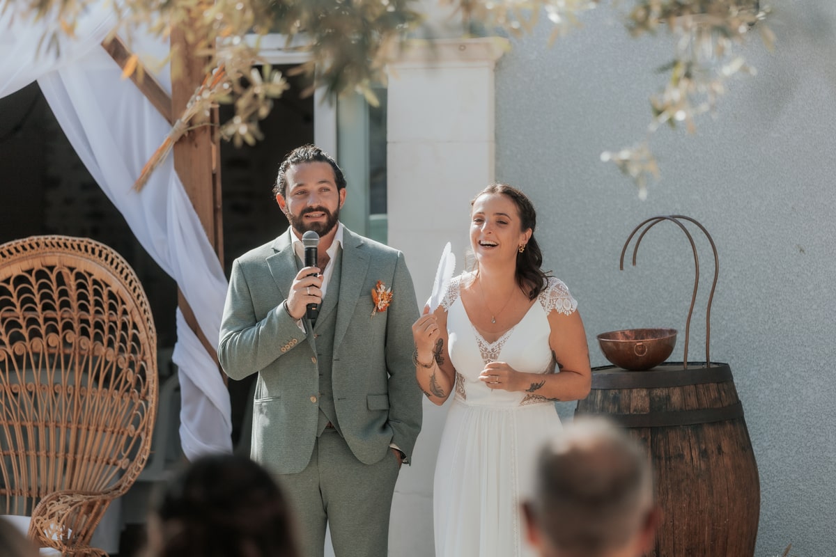 Couple de mariés souriants s'exprimant au micro lors d'une cérémonie en plein air à la Villa des 7 Moulins.