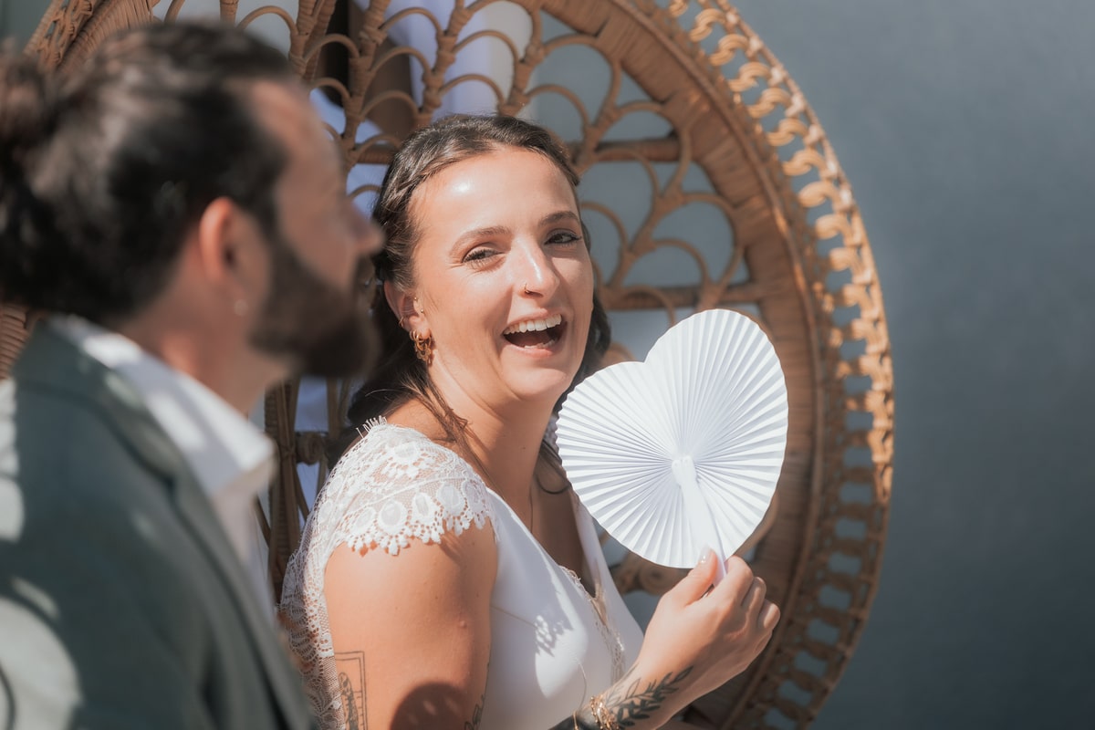 Mariée souriante tenant un éventail en forme de cœur, assise aux côtés de son marié à la Villa des 7 Moulins.