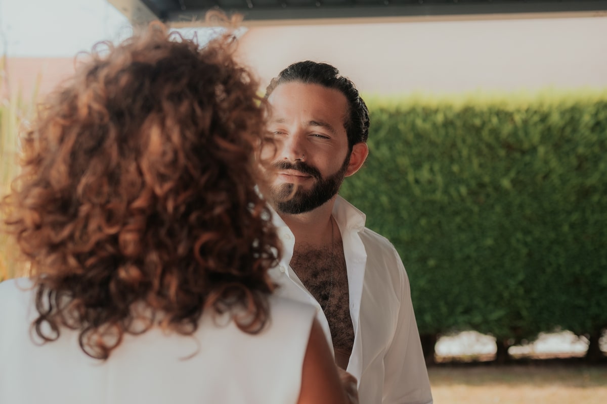 Homme souriant dans une chemise blanche, regardant une femme aux cheveux bouclés dans un jardin verdoyant.