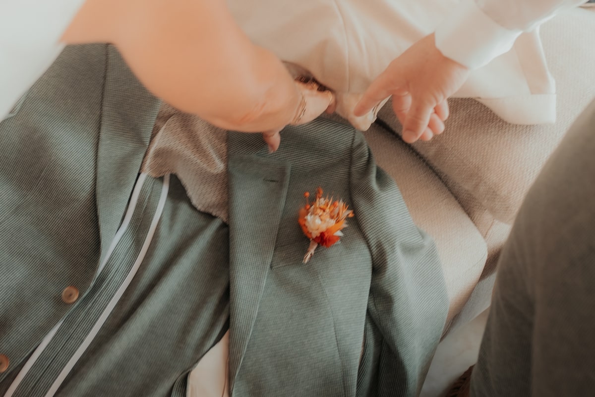 Des mains ajustent une veste élégante avec une boutonnière orange, préparatifs du mariage à la Villa des 7 Moulins.