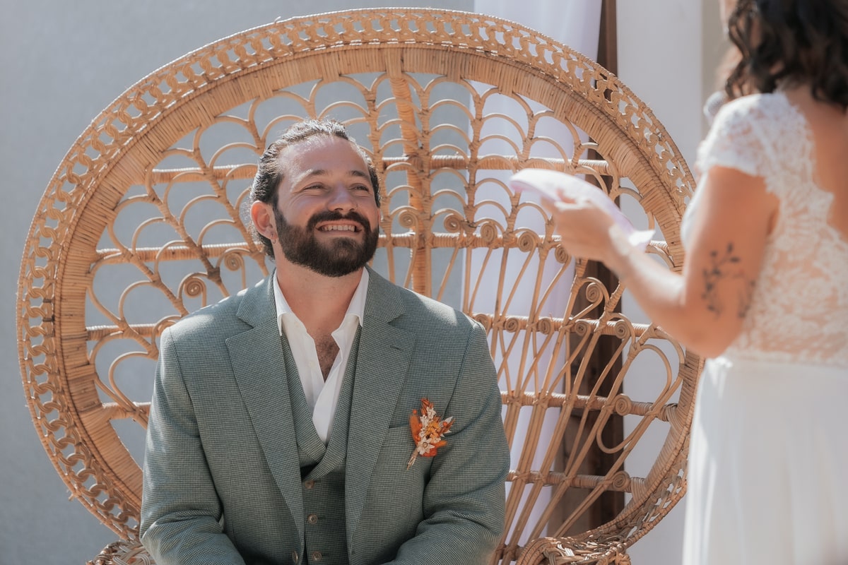 Marié souriant écoutant son épouse devant une chaise en rotin lors du mariage à la Villa des 7 Moulins.